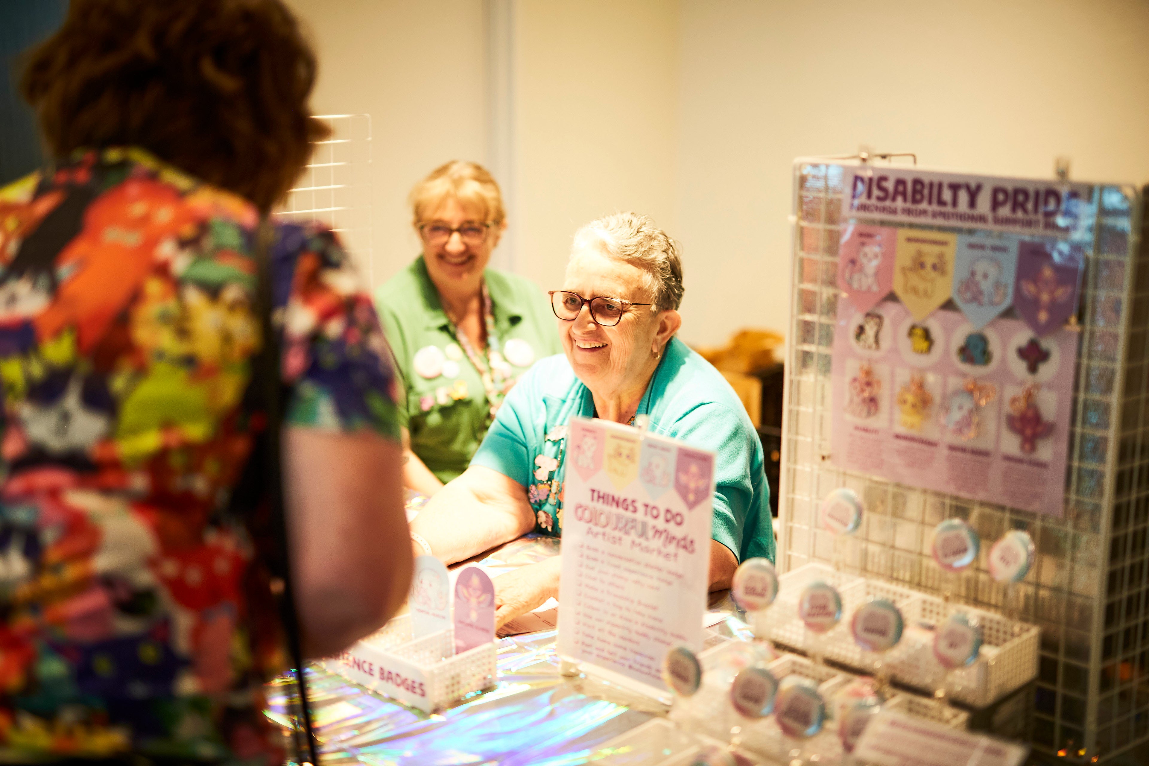Woman with glasses interacting with another person at a table with various items, including a Disability Pride poster.