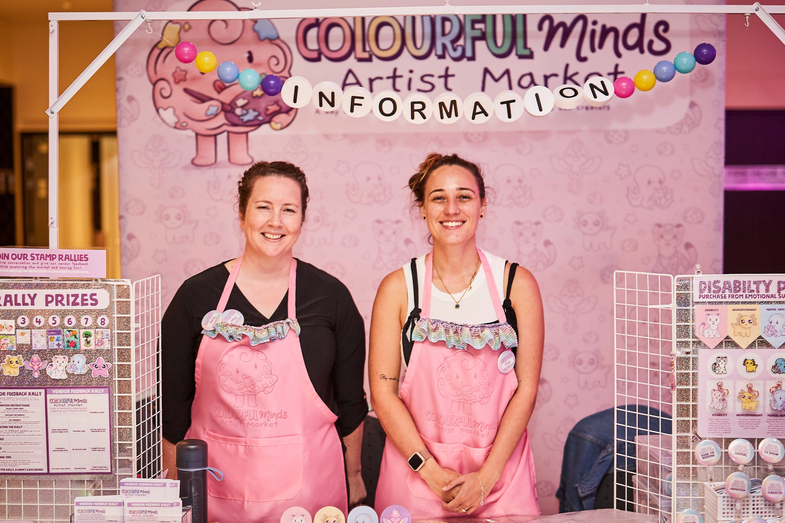 Two individuals in pink aprons standing behind a table with promotional materials at the Colourful Minds Artist Market.
