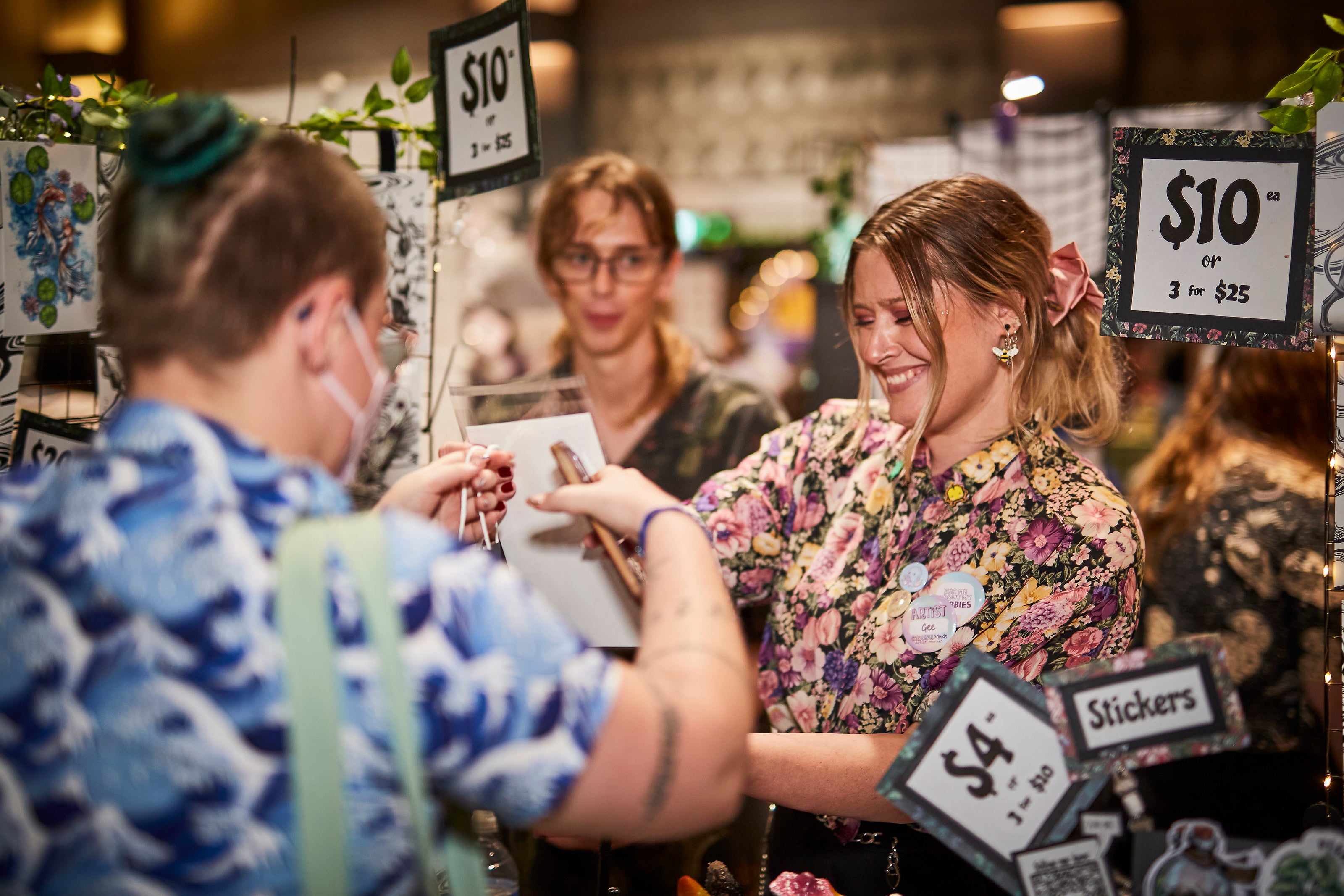 Two women interacting at a craft fair with booth signs displaying prices and items for sale.